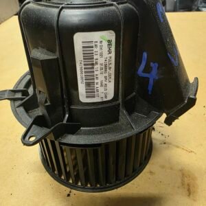Close-up of a dusty black blower motor housing with a barcode label, mounted mounting tabs, and blue marker on a workshop bench.