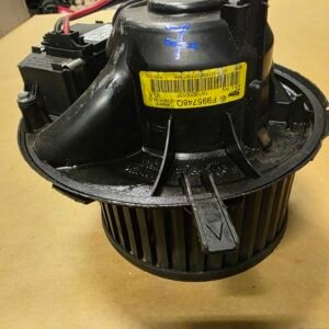 Close-up of a black circular automotive blower motor with a yellow barcode label and red wires in a workshop setting