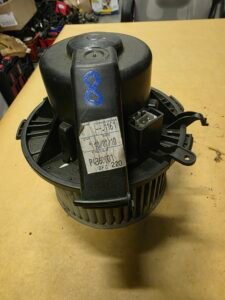 Black cylindrical automotive blower motor with a white label and a blue handwritten 8 on the side, on a wooden workbench.