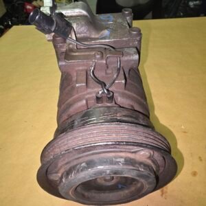 Rusty automotive pump with a large ribbed pulley and a black electrical connector on a yellow workbench surface in a cluttered workshop.