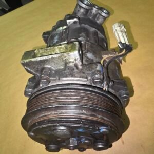 Old, dirty automotive air conditioning compressor with a large drive pulley and attached hoses on a brown surface.
