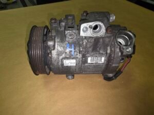 Automotive air conditioning compressor with a multi-groove pulley on the left and a weathered metal body on a brown surface, showing wiring at the bottom right.