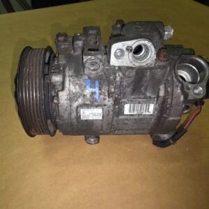Automotive air conditioning compressor with a multi-groove pulley on the left and a weathered metal body on a brown surface, showing wiring at the bottom right.